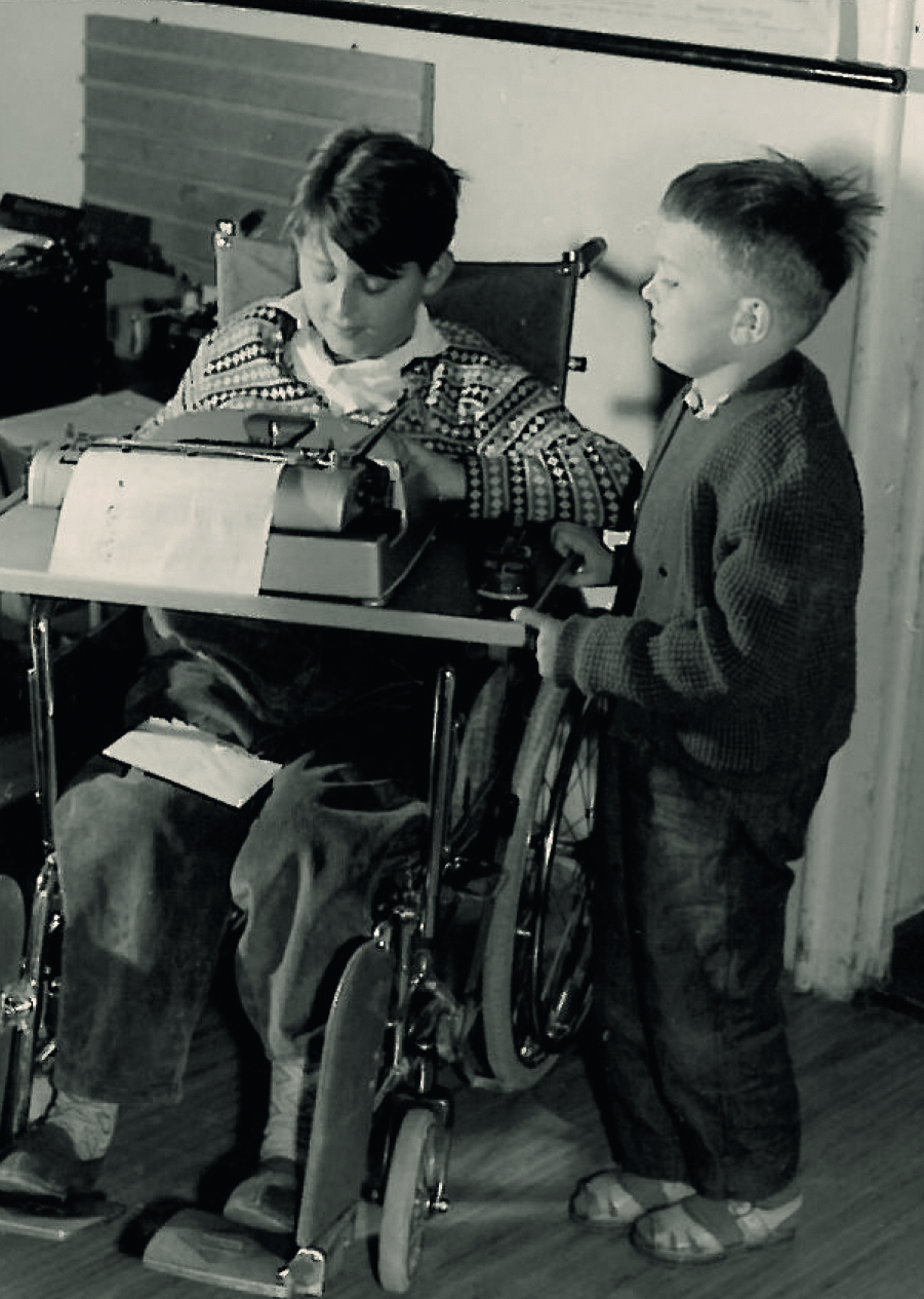 Zwei Schüler an einem Tisch vor einer Schreibmaschine, einer davon sitzt im Rollstuhk