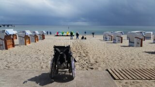Rollstuhl am Strand, davor stehen Strandkörbe