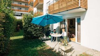 Zwei Personen sitzen auf Terrasse vor ihrer Wohnung mit Sonnenschirm