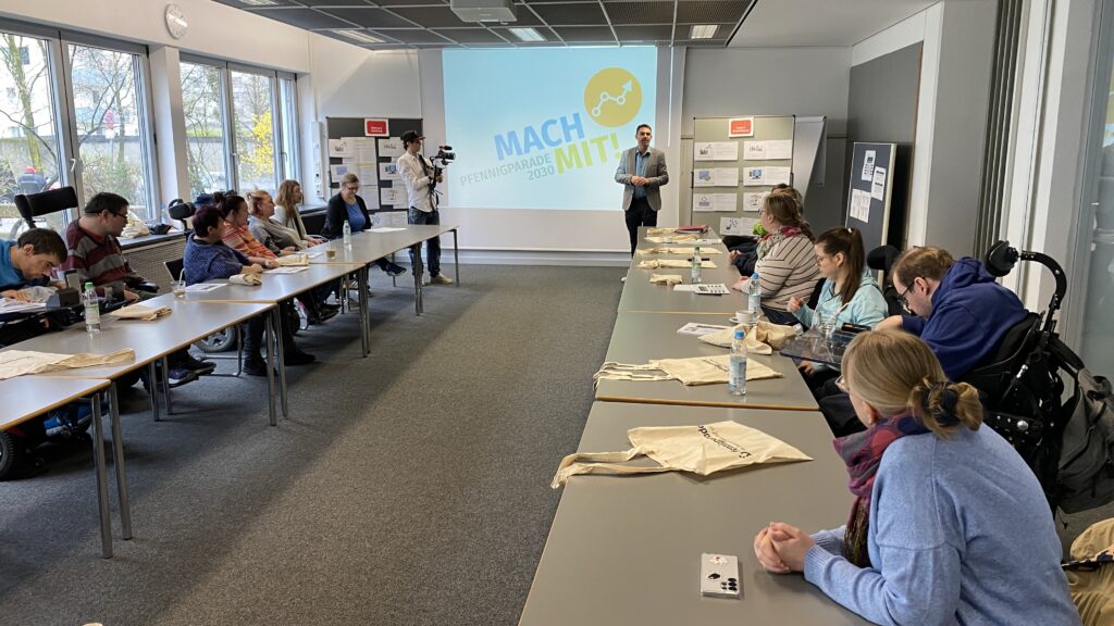 Viele Menschen mit und ohne Behinderung in Konferenzraum vor Leinwand Text: Pfennigparade 2030 Mach mit